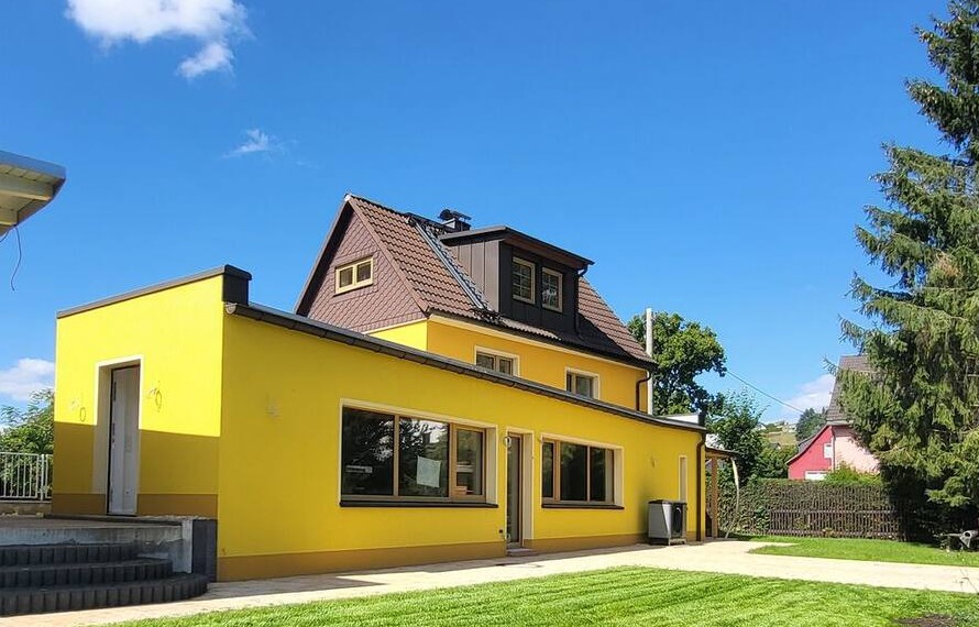 Das gelbe Haus mit hellem Anbau und Eingang, oben die Dachgaube, davor Rasen