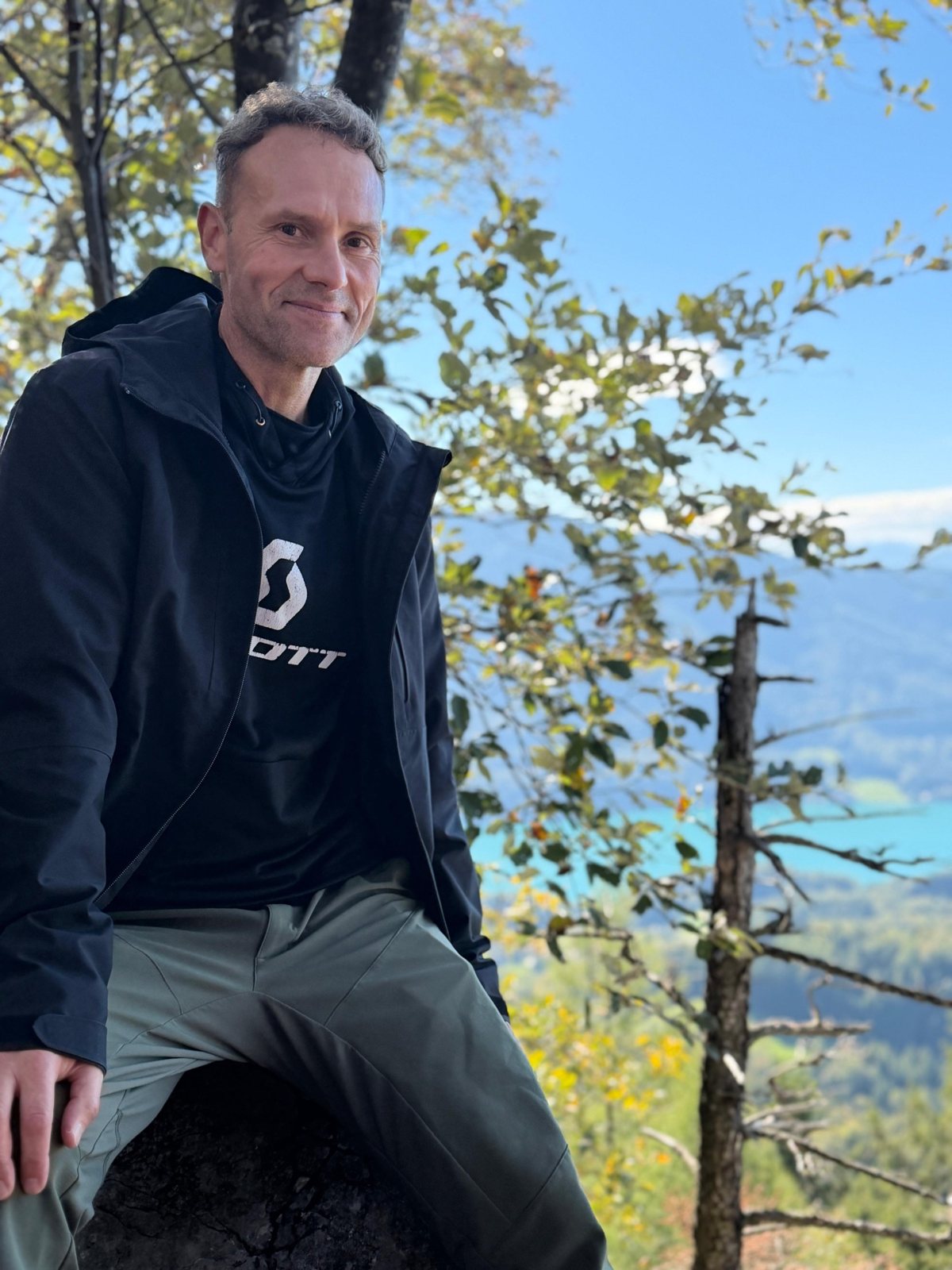 Roman, der Gastgeber, sitzt lächelnd auf einem Felsen im Wald, hinter ihm Bäume und ein See im Tal