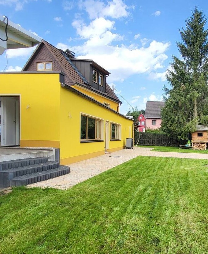 Das gelbe Haus mit hellem Anbau, Treppe zur Terrasse, Rasen und hinten das Holzspielhaus